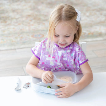 A young blonde child in a purple bib sits at a white table, removing a lid from Bumkins Silicone Grip Tray: Marble with utensils nearby.