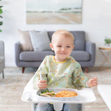 A toddler in a high chair enjoys food from a Bumkins Silicone Grip Tray: Marble. A gray couch and painting add charm to the cozy scene.