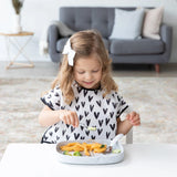 A girl in a heart-patterned bib enjoys a meal from a Bumkins Silicone Grip Tray: Marble at a white table with a gray sofa behind her.