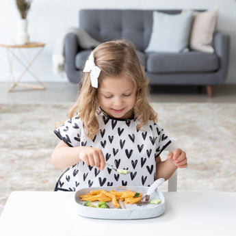 A girl in a heart-patterned bib enjoys a meal from a Bumkins Silicone Grip Tray: Marble at a white table with a gray sofa behind her.