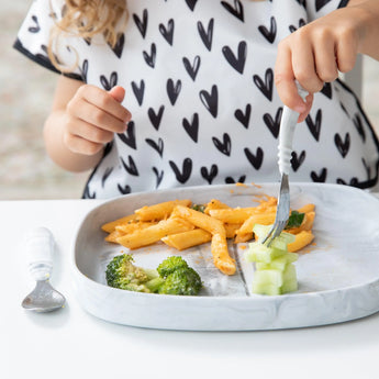 Close-up of a child enjoying pasta, broccoli, and cucumber from Bumkins Silicone Grip Tray: Marble, wearing a white bib with black hearts.