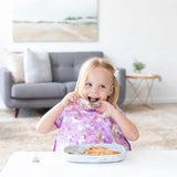 A little blonde girl in a purple bib happily eats from a Bumkins Silicone Grip Tray: Marble with a spoon at a white table.