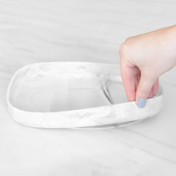 A hand holds a Bumkins Silicone Grip Tray: Marble, demonstrating the suction cup bottom on a marble surface.