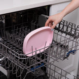 A hand places a Bumkins Silicone Grip Tray: Pink into the top rack of an open dishwasher.