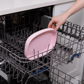 A hand places a Bumkins Silicone Grip Tray: Pink into the top rack of an open dishwasher.
