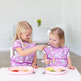 Two blonde kids in purple bibs enjoy meals at a white table; one uses a spoon to feed the other from a Bumkins Silicone Grip Tray: Pink.