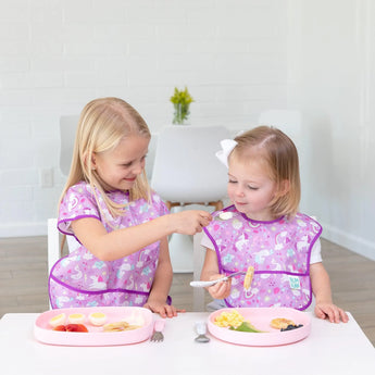 Two blonde kids in purple bibs enjoy meals at a white table; one uses a spoon to feed the other from a Bumkins Silicone Grip Tray: Pink.