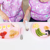 Two kids in unicorn bibs enjoy breakfast at a white table, with one eating from a Bumkins Silicone Grip Tray: Pink.