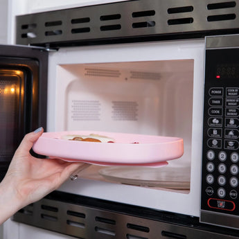 A hand places a Bumkins Silicone Grip Tray: Pink filled with food into an open microwave.
