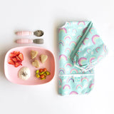 Bumkins Silicone Grip Tray: Pink with fruit and yogurt sits next to utensils, and a folded rainbow-patterned bib on a white background.