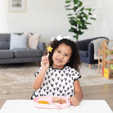A curly-haired child holds a fork with a star-shaped piece of fruit, with snacks on a Bumkins Silicone Grip Tray: Pink on a white table.