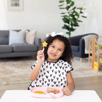 A curly-haired child holds a fork with a star-shaped piece of fruit, with snacks on a Bumkins Silicone Grip Tray: Pink on a white table.