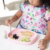 A child with a colorful bib sits at a table, with a Bumkins Silicone Grip Tray: Pink holding rolled sandwiches, cucumbers, and blueberries.
