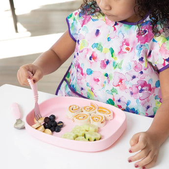 A child with a colorful bib sits at a table, with a Bumkins Silicone Grip Tray: Pink holding rolled sandwiches, cucumbers, and blueberries.