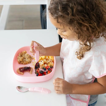 A child in a pink bib uses a fork to eat berries and oatmeal from Bumkins Silicone Grip Tray: Pink at a white table.