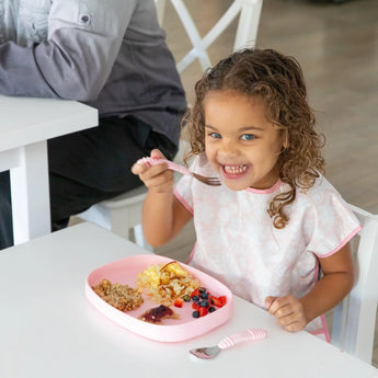 A girl with curly hair enjoys breakfast from a Bumkins Silicone Grip Tray: Pink filled with eggs, berries, and toast at a white table.