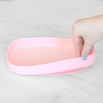 A hand holds a Bumkins Silicone Grip Tray: Pink, demonstrating the suction cup bottom on a marble surface.