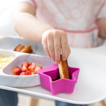Close-up of a child dipping French toast in a fuchsia Bumkins Silicone Little Dipper 3-Pack: Lollipop, attached to a plate on a high chair.