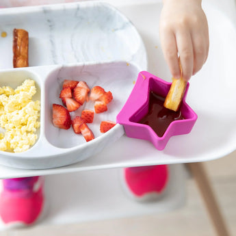 Close-up of a child in a high chair dipping bread in sauce from a fuchsia Bumkins Silicone Little Dipper 3-Pack: Lollipop next to a plate.