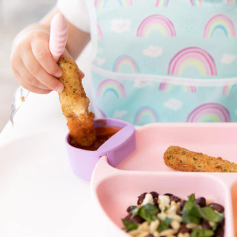 Close-up of a child dipping a mozzarella stick into sauce in a purple Bumkins Silicone Little Dipper 3-Pack: Lollipop next to a pink plate.