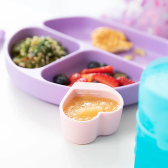 Close-up of a purple divided plate with salad and fruit, with a pink Bumkins Silicone Little Dipper 3-Pack: Lollipop with applesauce.