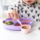 A child dips a spoon in a pink Bumkins Silicone Little Dipper 3-Pack: Lollipop attached to a purple sectioned plate of food at a table.