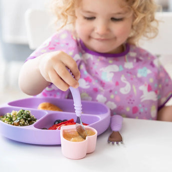 A curly-haired toddler in a purple bib eats applesauce from a pink Bumkins Silicone Little Dipper 3-Pack: Lollipop with a spoon.