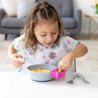 A child eats cereal from a gray bowl with a fuchsia Bumkins Silicone Little Dipper 3-Pack: Lollipop attached at a white table.