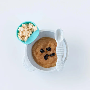 A bowl of applesauce sits on a white background, with a blue Bumkins Silicone Little Dipper Round 3-Pack: Gumdrop filled with cereal.
