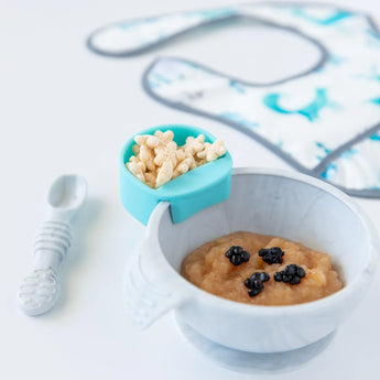 A blue Bumkins Silicone Little Dipper Round 3-Pack: Gumdrop with cereal attached to a bowl, with a spoon and bib nearby on a white background.