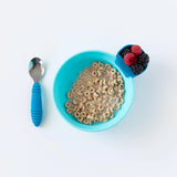 A blue cereal bowl with blue Bumkins Silicone Little Dipper Round 3-Pack: Gumdrop with berries sits next to a spoon on a white background.