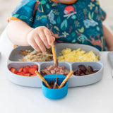 A child dipping pretzels into chocolate from a blue Bumkins Silicone Little Dipper Round 3-Pack: Gumdrop, attached to a plate of food.