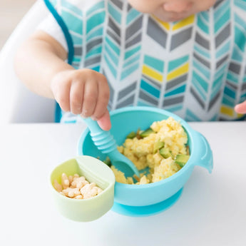 A baby enjoying scrambled eggs from a bowl, with a green Bumkins Silicone Little Dipper Round 3-Pack: Gumdrop with cereal attached.