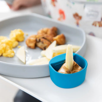 A blue Bumkins Silicone Little Dipper Round 3-Pack: Gumdrop with peanut butter next to a plate of apple slices, eggs, & chicken bites.