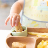 Close-up of a child's hand dipping a nugget into sauce in a green Bumkins Silicone Little Dipper Round 3-Pack: Gumdrop on a brown plate.