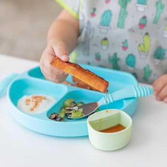 A child dips French toast into syrup in a green Bumkins Silicone Little Dipper Round 3-Pack: Gumdrop attached to a blue plate with food.
