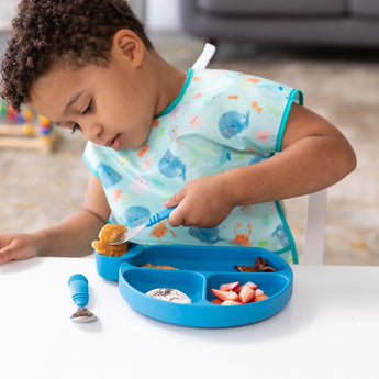 A child in a bib dips food in a blue Bumkins Silicone Little Dipper Round 3-Pack: Gumdrop attached to a blue dish using a fork at a table.