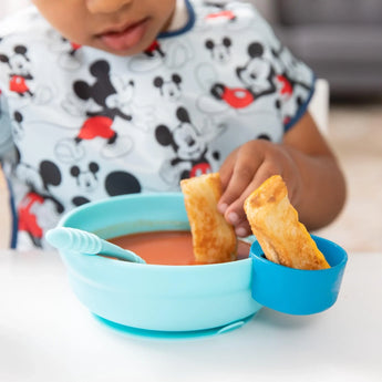 A child in a bib happily dips breadsticks from a blue Bumkins Silicone Little Dipper Round 3-Pack: Gumdrop attached to a bowl of soup.