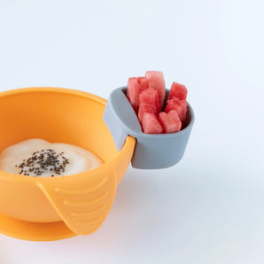 An orange bowl with yogurt, with a gray Bumkins Silicone Little Dipper Round 3-Pack: Taffy holding watermelon on a white background.