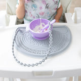 A child in a highchair enjoys yogurt from a purple bowl on a Bumkins Silicone Sensory Placemat: Gray, with a spoon tethered to the tray.