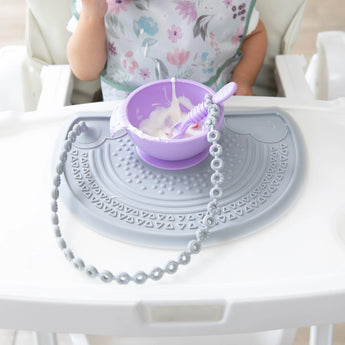A child in a highchair enjoys yogurt from a purple bowl on a Bumkins Silicone Sensory Placemat: Gray, with a spoon tethered to the tray.