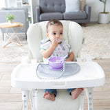 A baby in a high chair wearing a floral bib, using a purple bowl and spoon on a Bumkins Silicone Sensory Placemat: Gray.