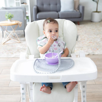 A baby in a high chair wearing a floral bib, using a purple bowl and spoon on a Bumkins Silicone Sensory Placemat: Gray.