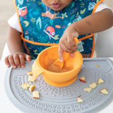 A toddler in a colorful bib eats with a spoon from an orange bowl on a Bumkins Silicone Sensory Placemat: Gray covered in diced fruit.