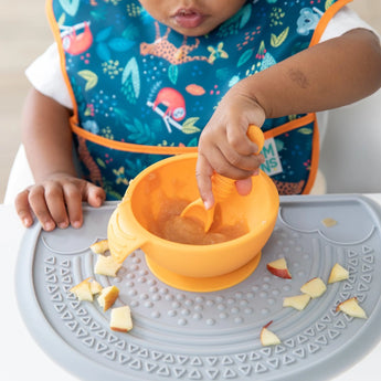 A toddler in a colorful bib eats with a spoon from an orange bowl on a Bumkins Silicone Sensory Placemat: Gray covered in diced fruit.