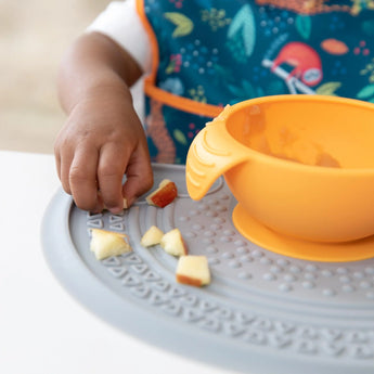 Close-up of a child at a table, picking up apple pieces from a Bumkins Silicone Sensory Placemat: Gray with an orange bowl on top.