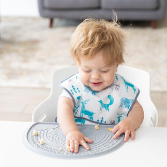 A curly-haired baby in a patterned bib plays with food on a Bumkins Silicone Sensory Placemat: Gray while seated in a high chair.