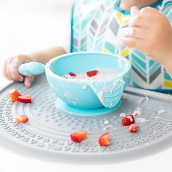 A child in a patterned bib spills strawberries and yogurt from a blue bowl onto a Bumkins Silicone Sensory Placemat: Gray.
