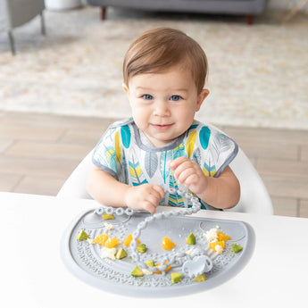 A toddler sits at a white table, enjoying fruit and avocado on a Bumkins Silicone Sensory Placemat: Gray.