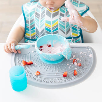 A baby messily eats yogurt from a blue bowl on a Bumkins Silicone Sensory Placemat: Gray, with a blue cup nearby.
